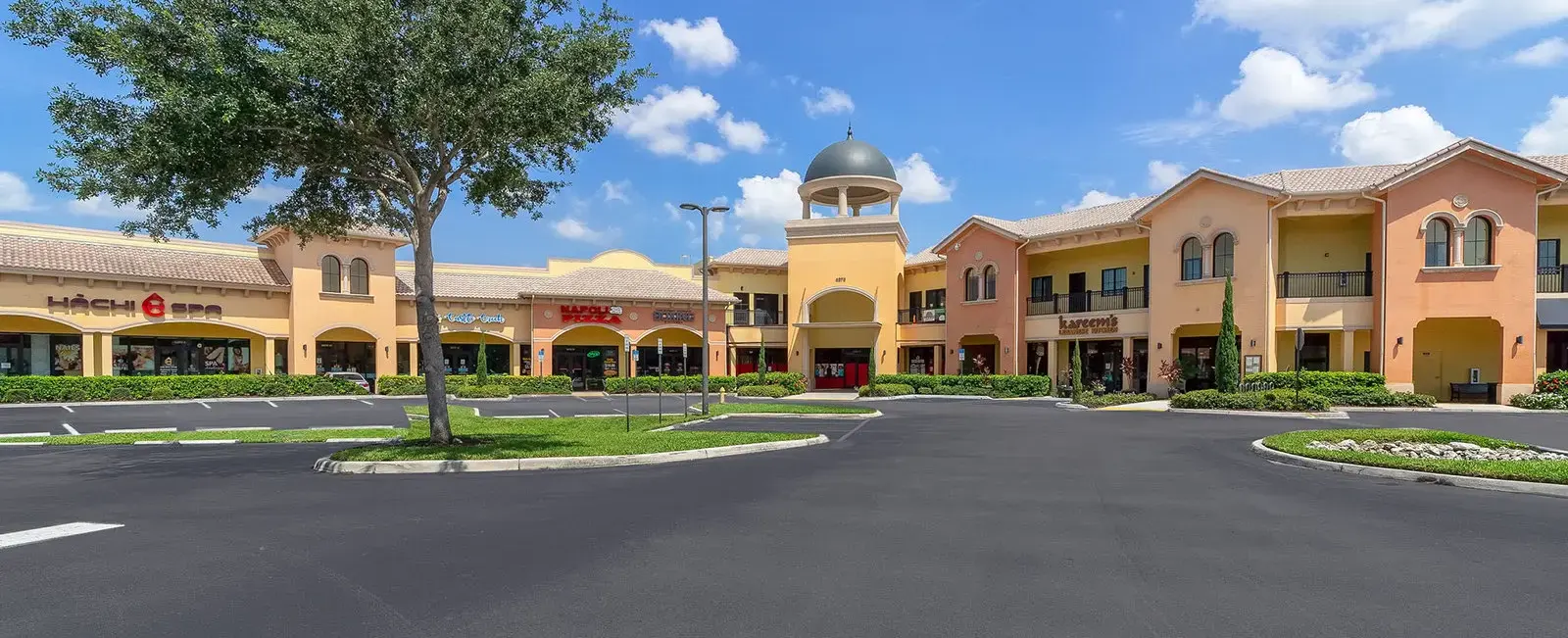 Front view of Sugden Park Plaza in Naples, Florida, featuring the commercial center's storefronts, customer entrances, covered walkways, landscaping, and parking area.