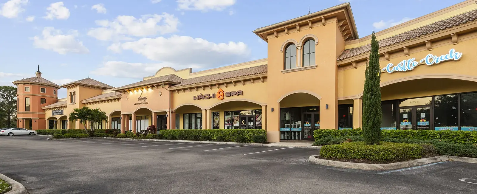 Close-up exterior view of a portion of Sugden Park Plaza in Naples, Florida, featuring storefronts, building architecture, signage, and pedestrian access areas.